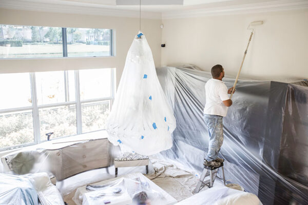 Painter's Plastic - Painter working with roller on walls in room where all furniture and carpet are covered in plastics